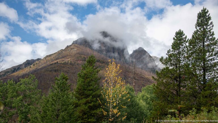 Jagged mountain peaks partly wrapped in low cloud above green conifers and a bright yellow autumn tree.