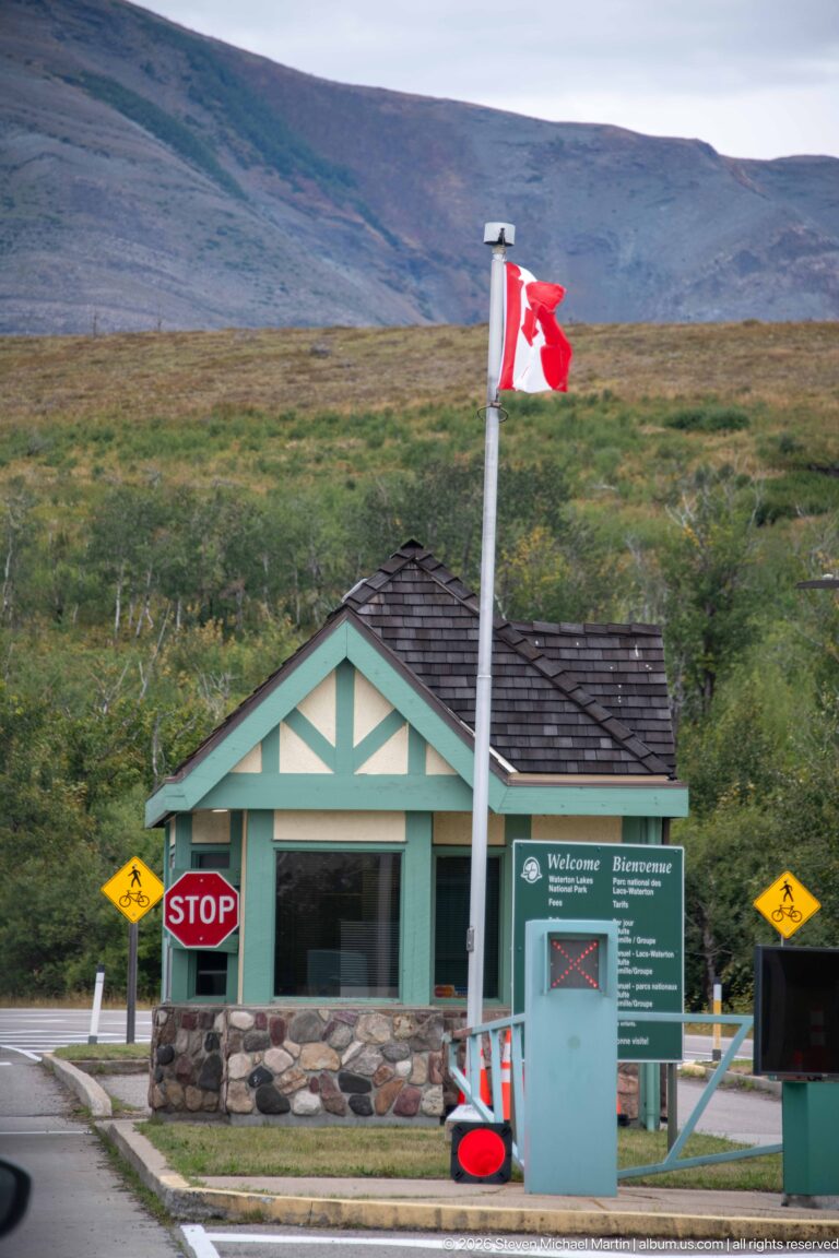 Green-and-cream park entrance building beneath a Canadian flag, with Welcome panel, stop sign, and wildlife-warning signs.