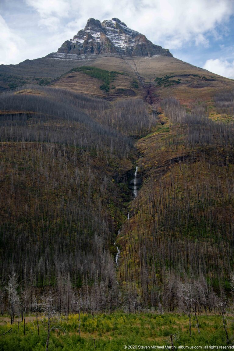 Broad mountain and burned forest filled with gray snags, crossed by a narrow cascade and bands of autumn-colored vegetation.