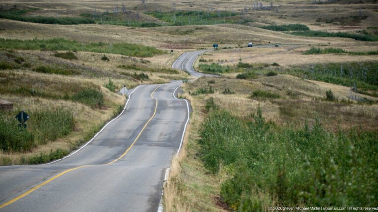Two-lane paved road curving and rising through open grassy foothills with low shrubs, roadside signs, and a few vehicles.