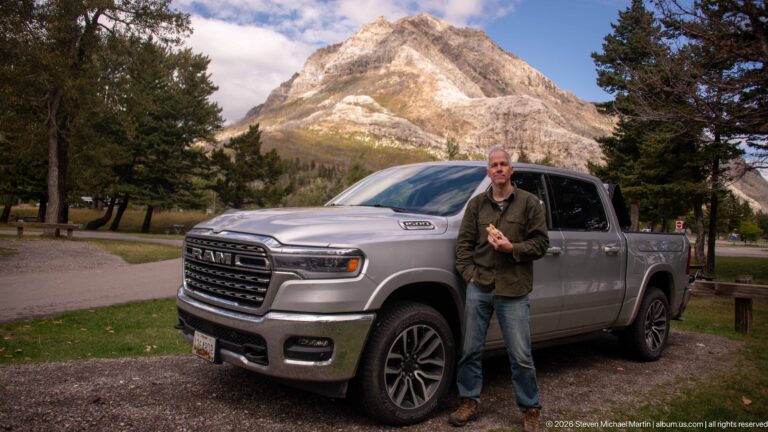 Visitor standing beside a silver RAM pickup truck at a campground, with a steep pale mountain and conifers behind.