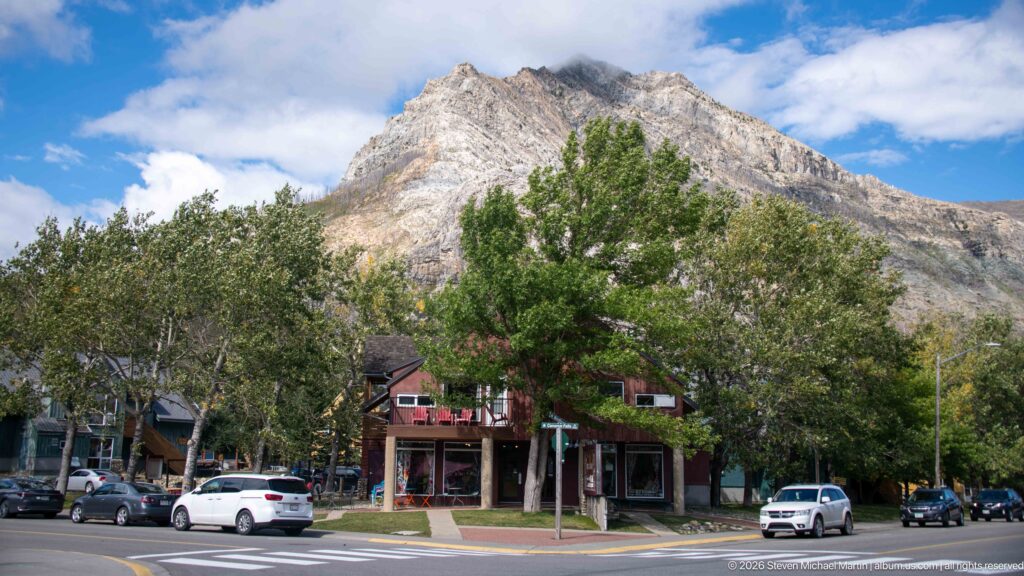 Waterton townsite street with low commercial buildings, leafy trees, cars, and a steep pale mountain rising directly behind.