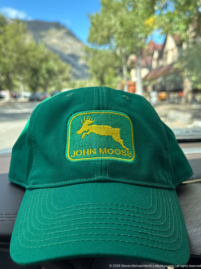 Green baseball cap on a vehicle dashboard embroidered with a yellow moose emblem and the words John Moose.