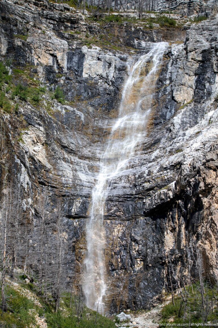 Broad waterfall divided into white ribbons over a dark horizontally layered cliff, with sparse dead trunks at the base.