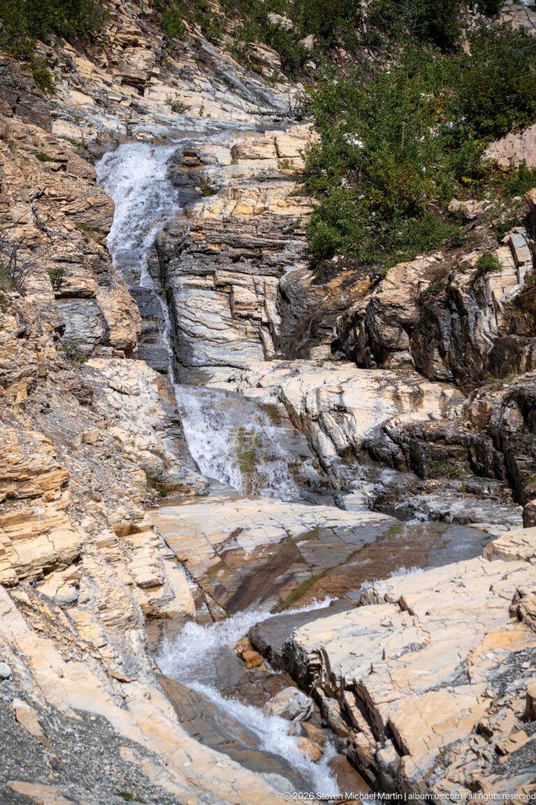 Mountain stream flowing through shallow steps and pools across broad pale layered bedrock, with low vegetation along the upper edge.
