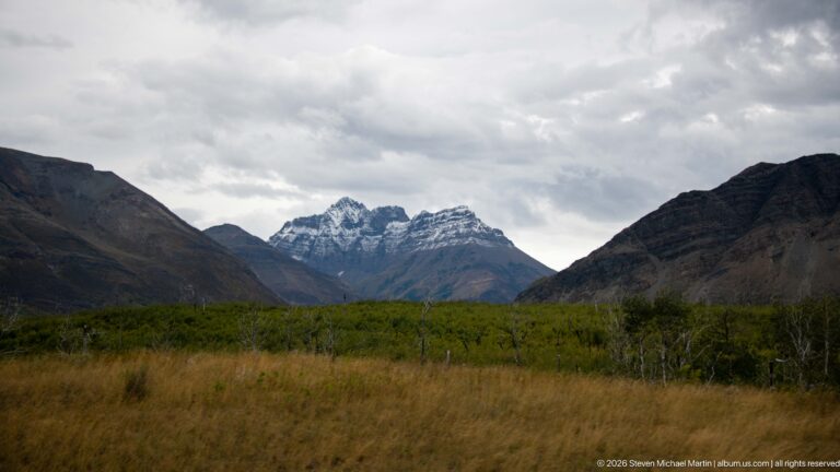 Jagged gray peaks at the head of a broad valley, framed by dark slopes above tawny grass and low shrubs.