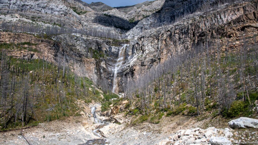Tall multi-tiered waterfall descending a layered cliff into a rocky basin with many standing dead trees and a stream below.