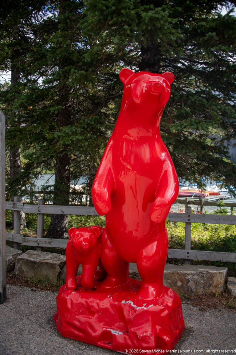Glossy bright-red sculpture of an upright adult bear with a smaller cub beside it, set among conifer trees.
