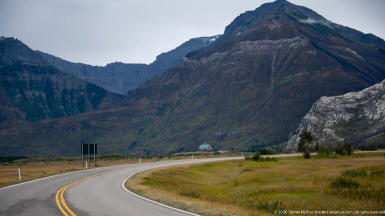 Curving two-lane road crossing open grassland toward steep dark mountains and a pale rock face, with a small building ahead.