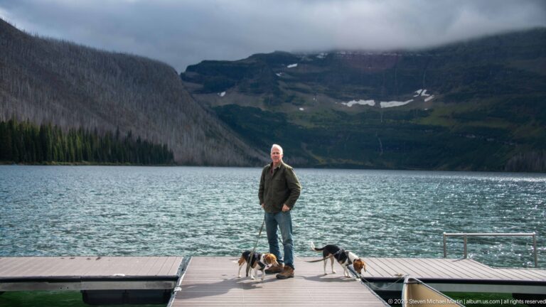 Visitor with two beagles on a Cameron Lake dock, one dog turned toward the water beneath cloud-covered mountain walls.
