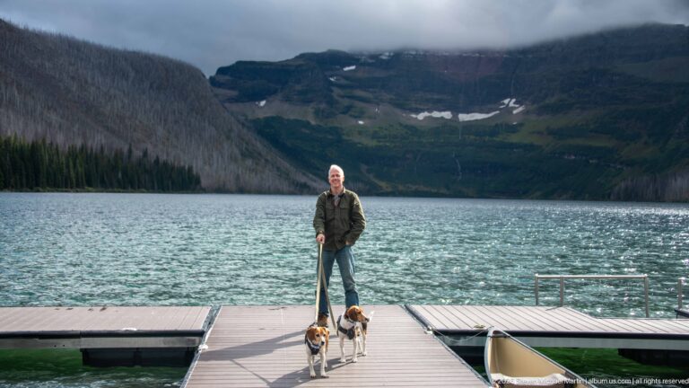 Visitor standing with two beagles on a floating Cameron Lake dock, with rippled water and cloud-covered mountains behind.