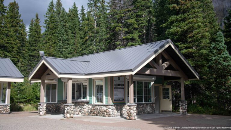 Gabled Cameron Lakeshore visitor building with timber entrance canopy, large windows, gray metal roof, and rounded-stone piers.