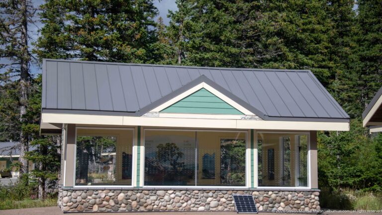 Small Cameron Lakeshore building with broad windows, green gable siding, gray metal roof, rounded-stone base, and small solar panel.