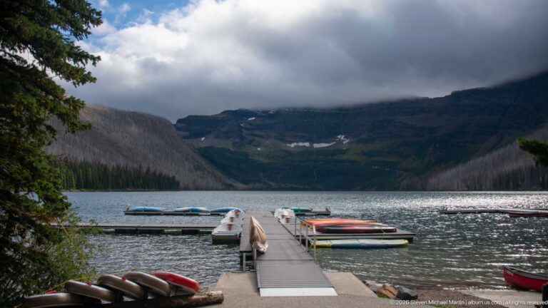 Floating dock extending into Cameron Lake with small boats moored alongside and cloud-covered mountains enclosing the far shore.