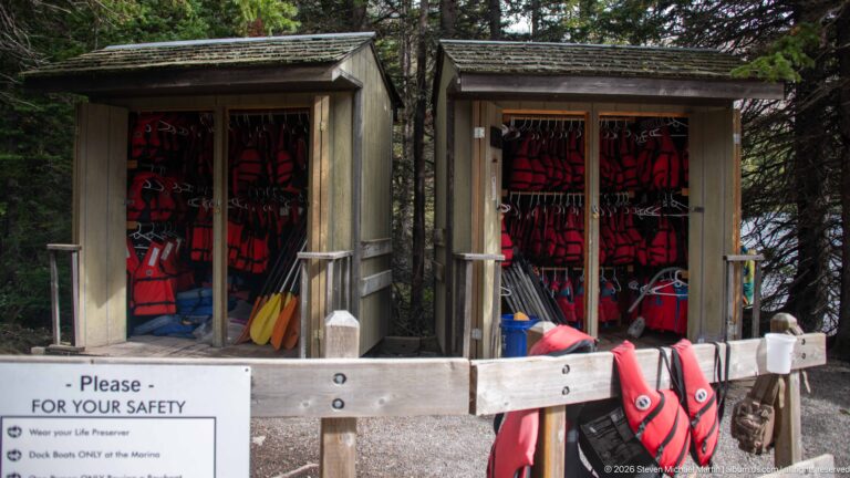 Open wooden equipment bays holding red life jackets, paddles, and boating gear near Cameron Lake, with a safety notice across the front.