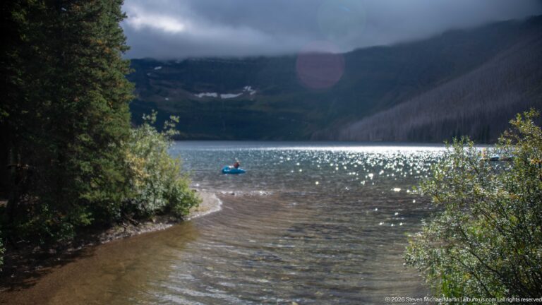 Single paddler in a small blue craft on sunlit Cameron Lake, framed by shoreline shrubs and cloud-covered mountain walls.
