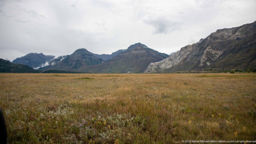 Wide tawny grassland stretching toward a continuous chain of dark rugged mountains beneath an overcast sky.
