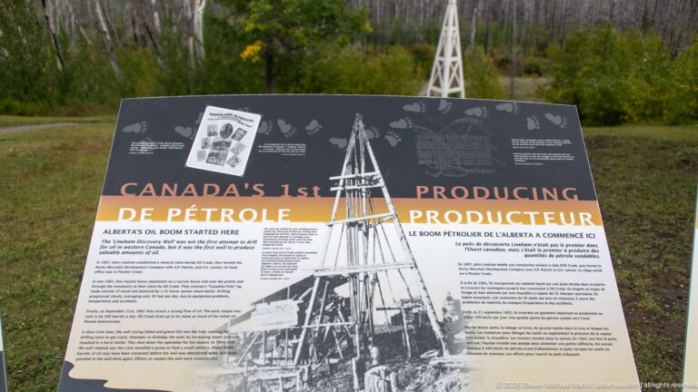Interpretive panel titled Canada’s 1st Producing Oil Well, with an archival derrick photograph and reconstructed derrick behind it.