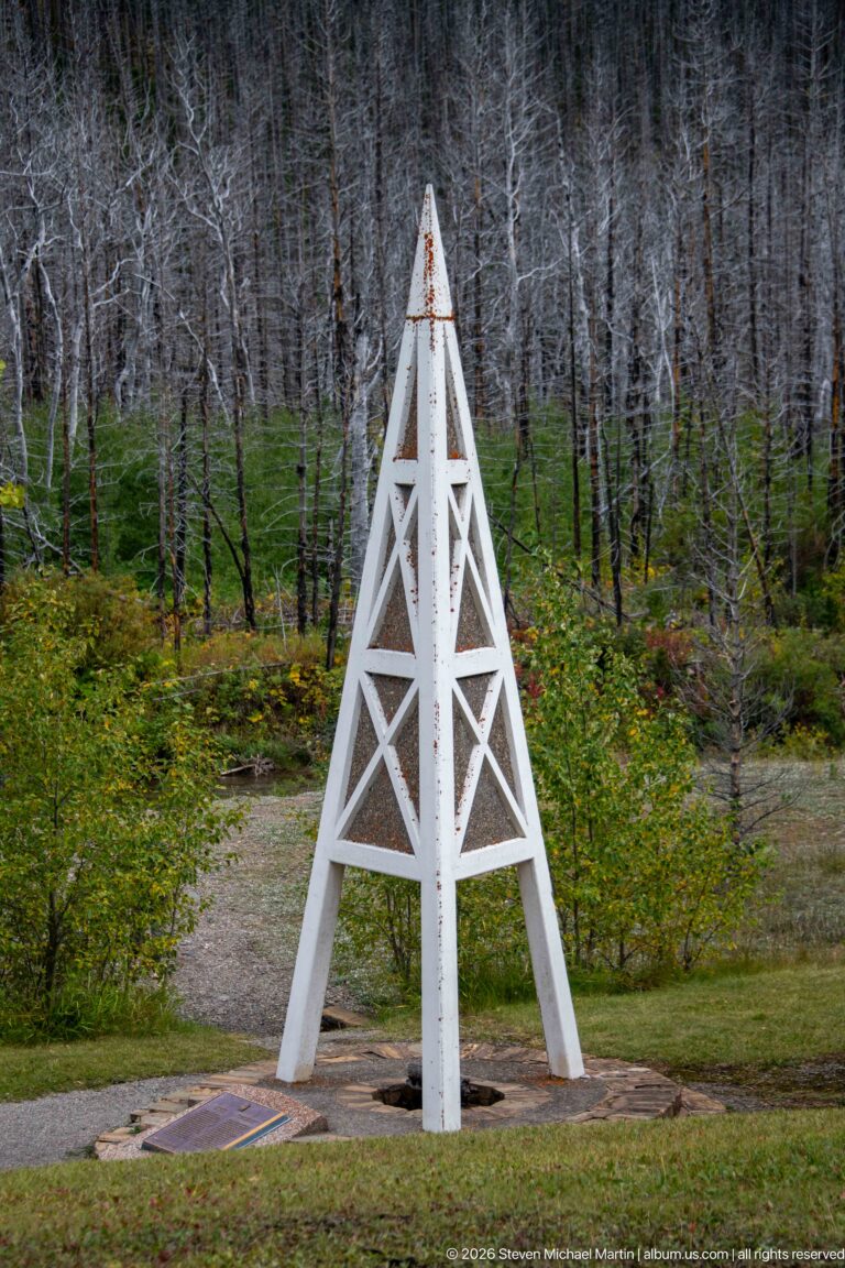 White timber oil derrick monument on a circular base amid green groundcover and numerous standing dead trees.