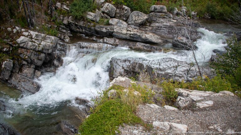 White water cascading over layered gray rock ledges around a large central boulder, bordered by low green vegetation.