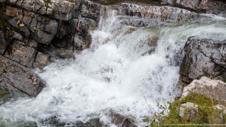 Fast white water cascading through a narrow channel of angular layered gray rock, with small ledges and turbulent pools.