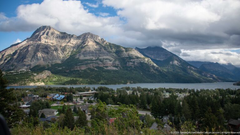 Waterton townsite roofs and trees below Upper Waterton Lake, backed by layered mountains beneath broken clouds.