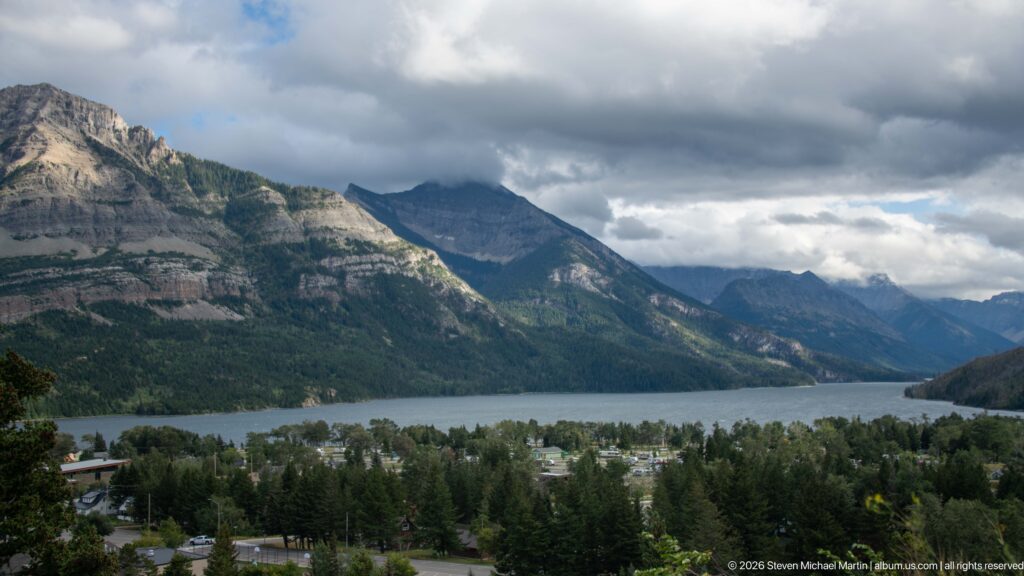 Waterton townsite among trees below Upper Waterton Lake, with steep mountains enclosing the valley under heavy clouds.