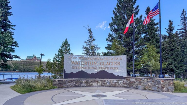 Waterton-Glacier International Peace Park monument beside Canadian and U.S. flags, with a circular pavement emblem and lakeshore beyond.