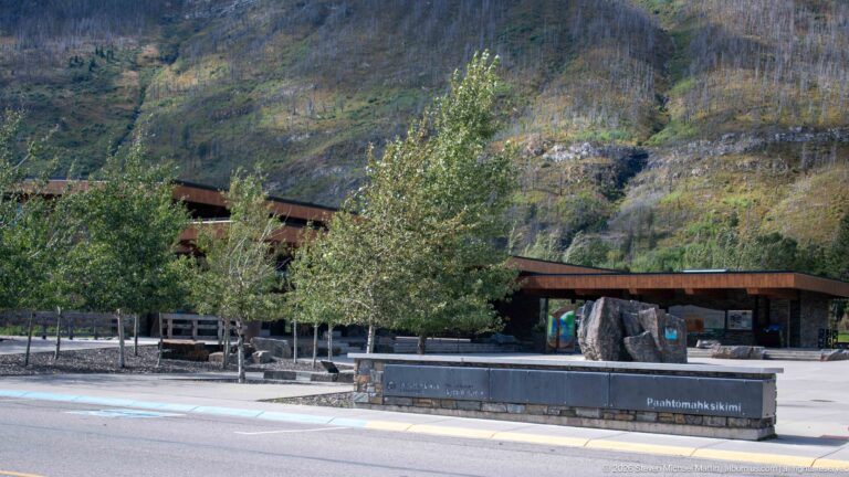 Visitor centre plaza with young trees, low timber buildings, stone sign, and steep mountain slope behind.