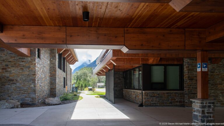 Covered visitor centre passage with heavy timber beams, wood ceiling, stone walls, and sunlit walkway ahead.