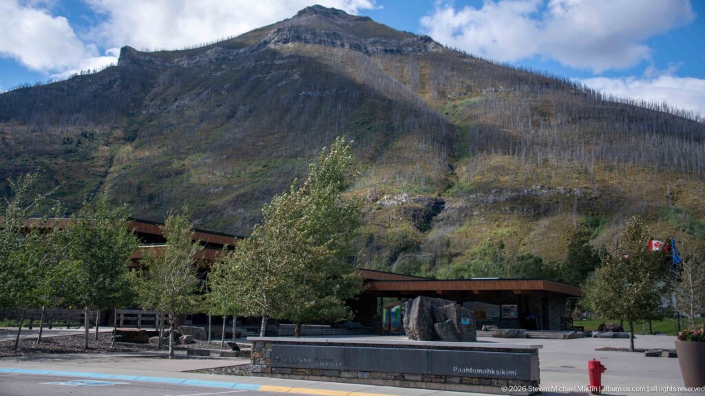 Low visitor centre buildings and trees beneath a steep mountain, with flags at the right side of the plaza.