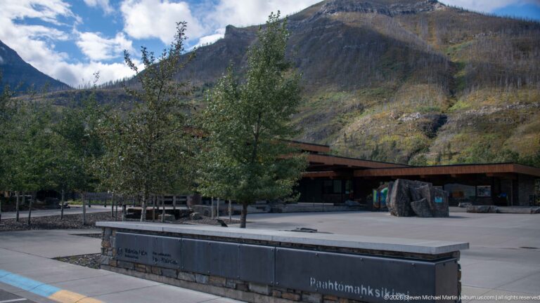 Paved visitor centre forecourt with trees, a boulder display, low timber roofs, and Paahtómahksikimi sign.