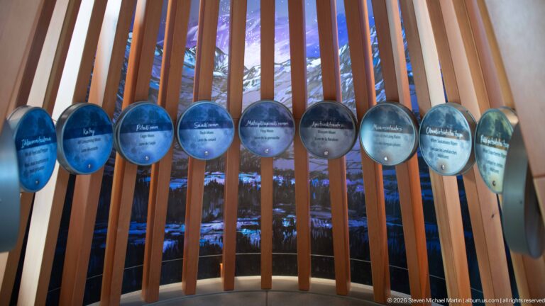 Row of bilingual circular interpretive discs mounted across vertical wooden slats, with mountains visible through gaps.