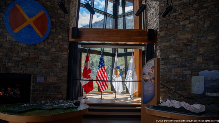 Stone-and-timber visitor center interior with four standing flags framed by tall windows and exhibit displays.