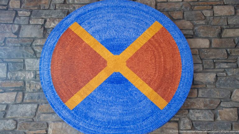 Large circular blue and rust fiber artwork with yellow X-shaped bands mounted on a stone wall.