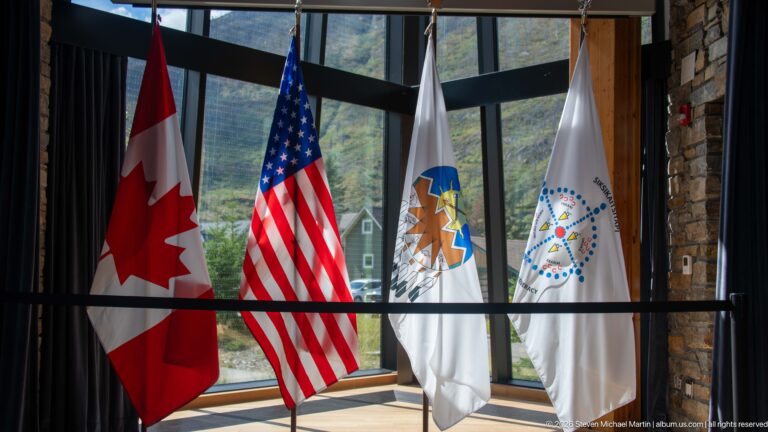 Canadian and United States flags beside two white emblem flags in front of tall visitor center windows.