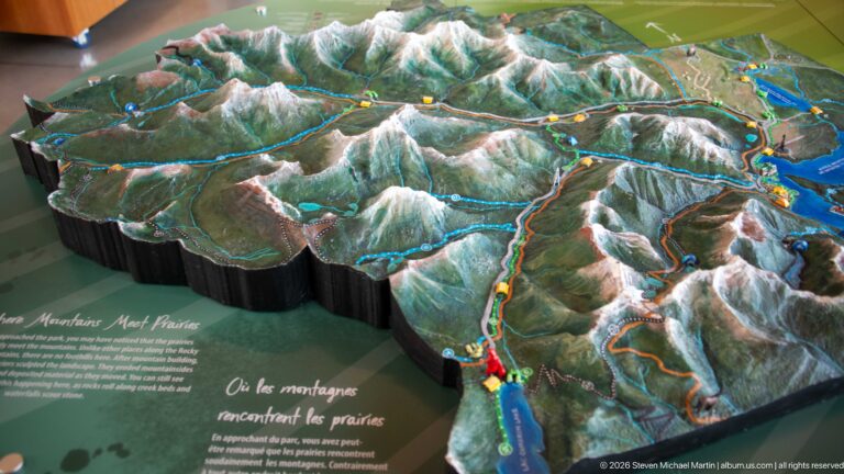 Oblique view of a three-dimensional relief map showing mountains, lakes, roads, and colored routes around Waterton.