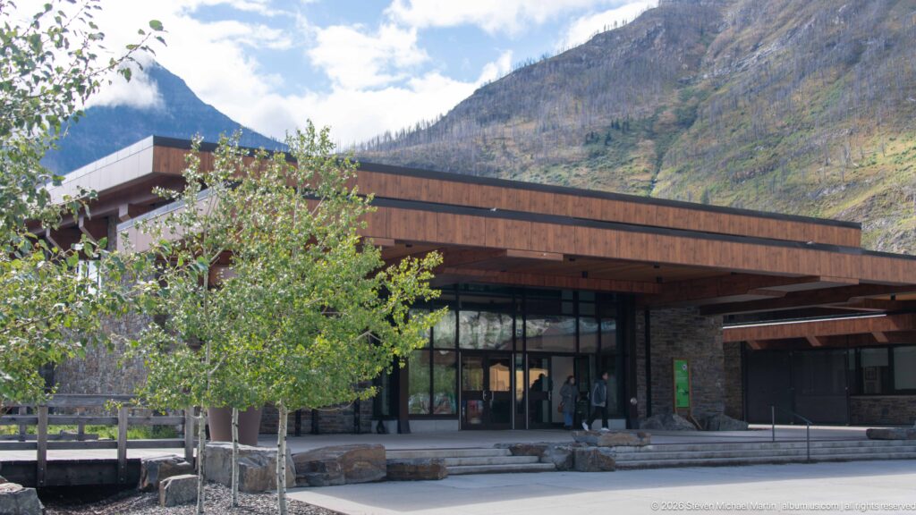 Glazed visitor centre entrance beneath a broad timber canopy, with stone walls, boulders, tree, and visitors.