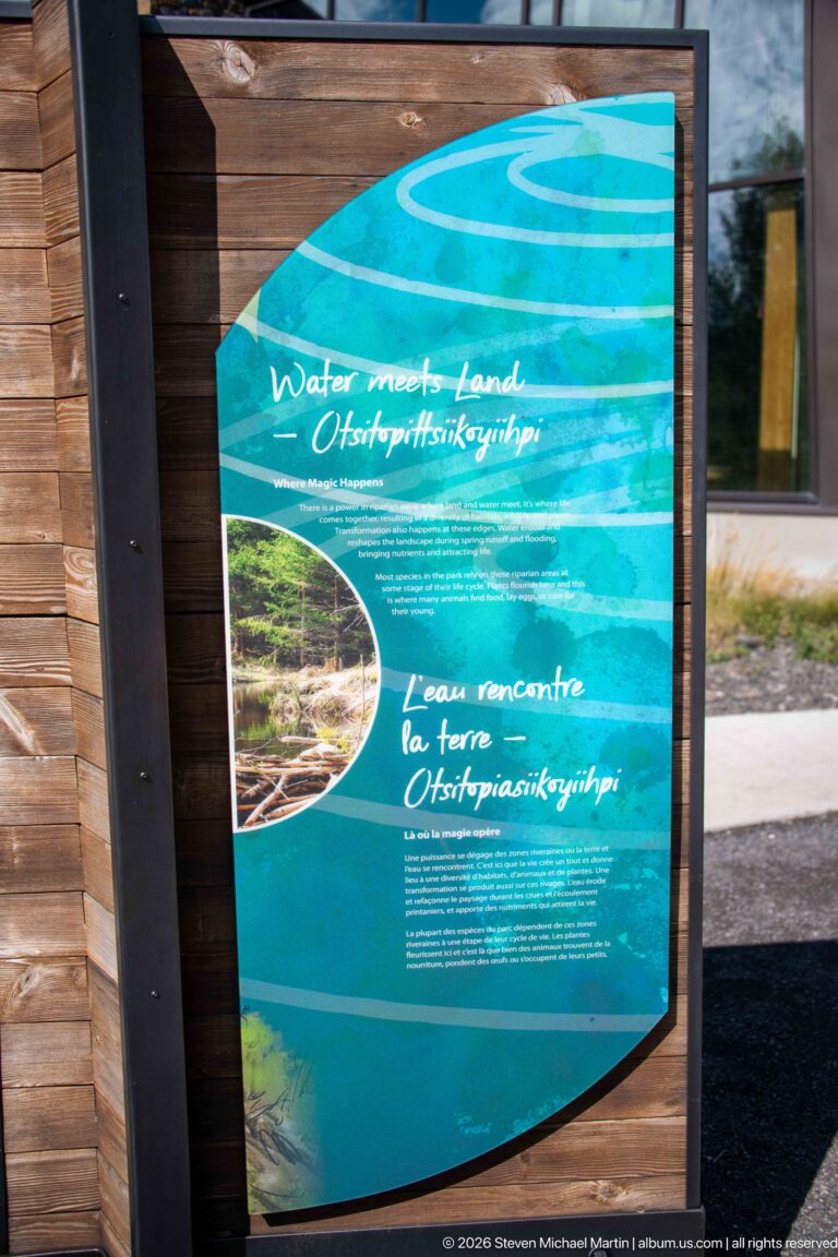 Turquoise outdoor panel titled Water meets Land with bilingual text and a circular landscape photograph in a wood frame.