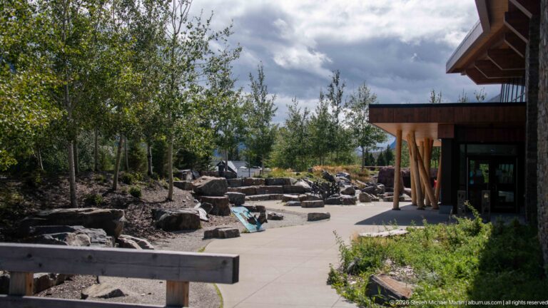 Paved visitor center courtyard with boulders, young trees, planted beds, and a timber-covered building entrance.