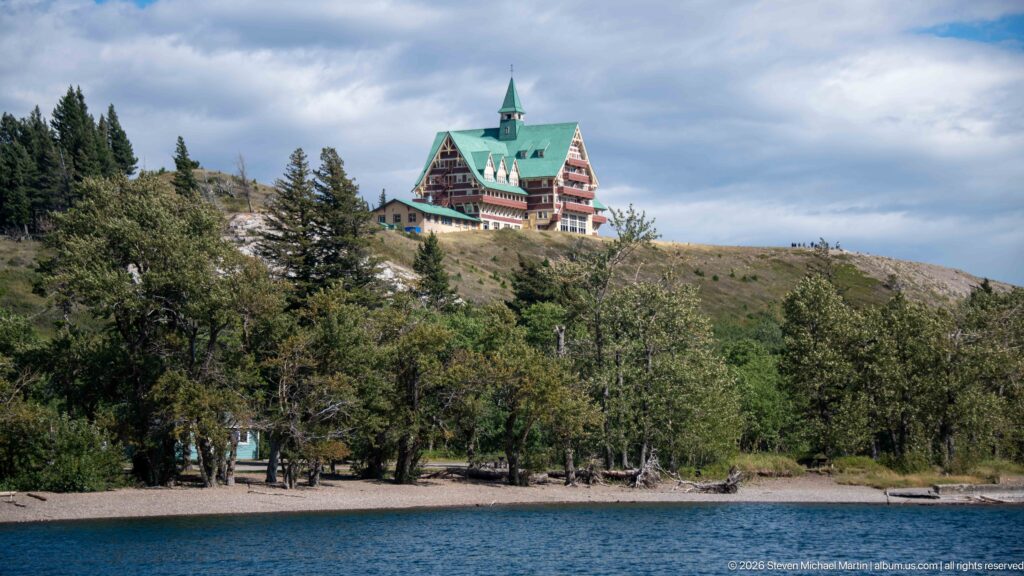 Prince of Wales Hotel with green roof and central tower standing on a bluff above blue lake water.