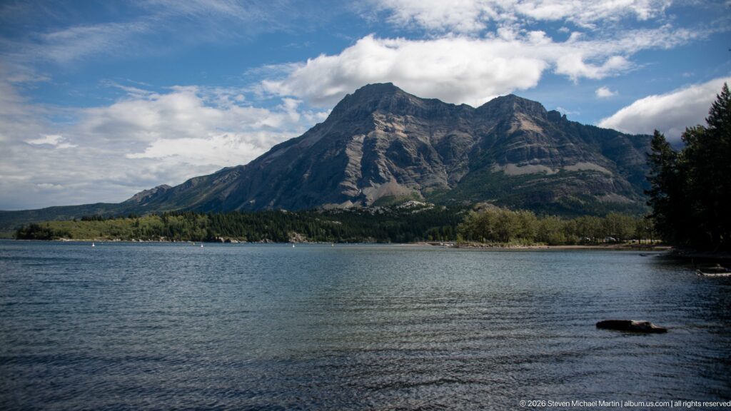 Broad gray mountain massif rising above a forested shoreline across calm blue lake water under broken clouds.