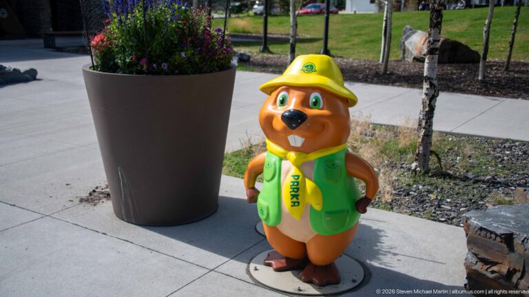 Beaver mascot figure wearing yellow ranger hat and green Parks vest beside a large outdoor planter.