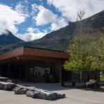 Timber visitor centre wing with boulders, Canadian and U.S. flags, trees, mountains, and clouds.