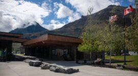 Timber visitor centre wing with boulders, Canadian and U.S. flags, trees, mountains, and clouds.