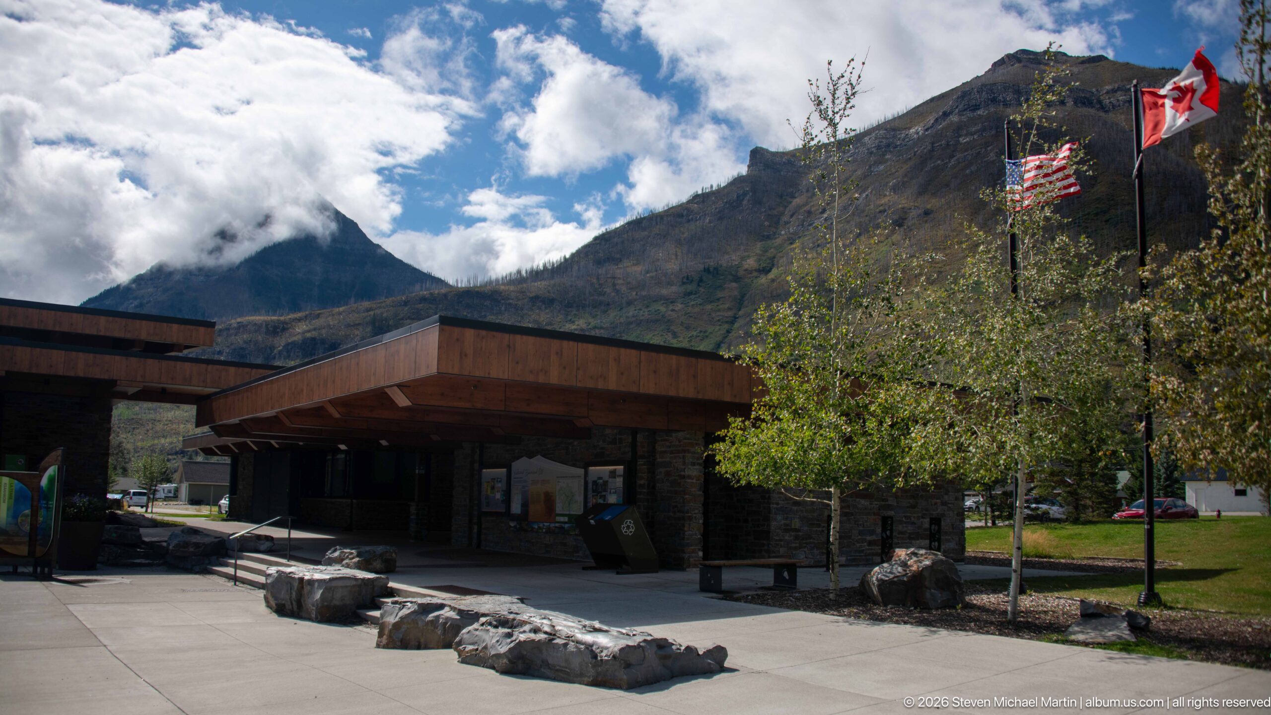 Timber visitor centre wing with boulders, Canadian and U.S. flags, trees, mountains, and clouds.