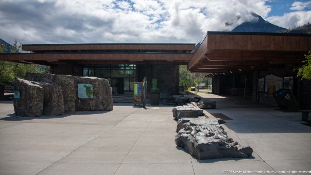 Open visitor centre plaza with interpretive boulder, low rock edging, timber roofs, and glazed entrance.
