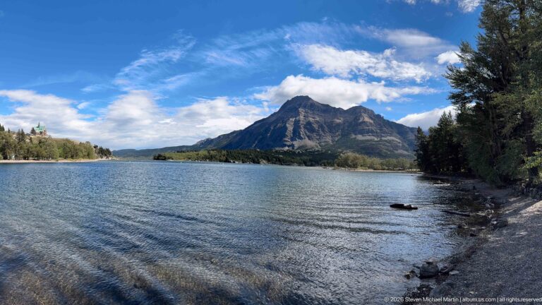 Wide view across rippled Upper Waterton Lake toward a rugged mountain, with trees framing both shores.
