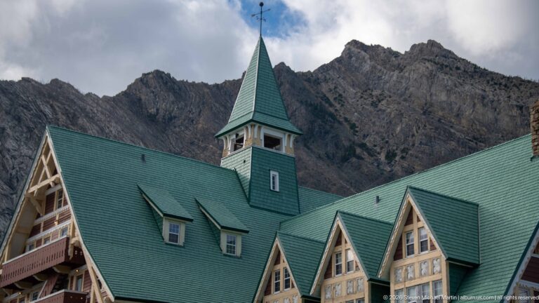Green roof and central roof feature of Prince of Wales Hotel with three projecting gables against a rocky mountain.