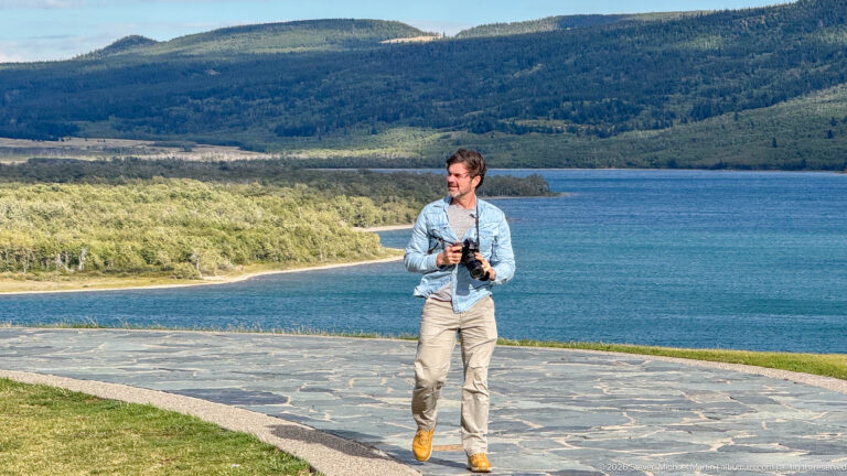 Person carrying a camera on a stone path, with Upper Waterton Lake and wooded shoreline behind.